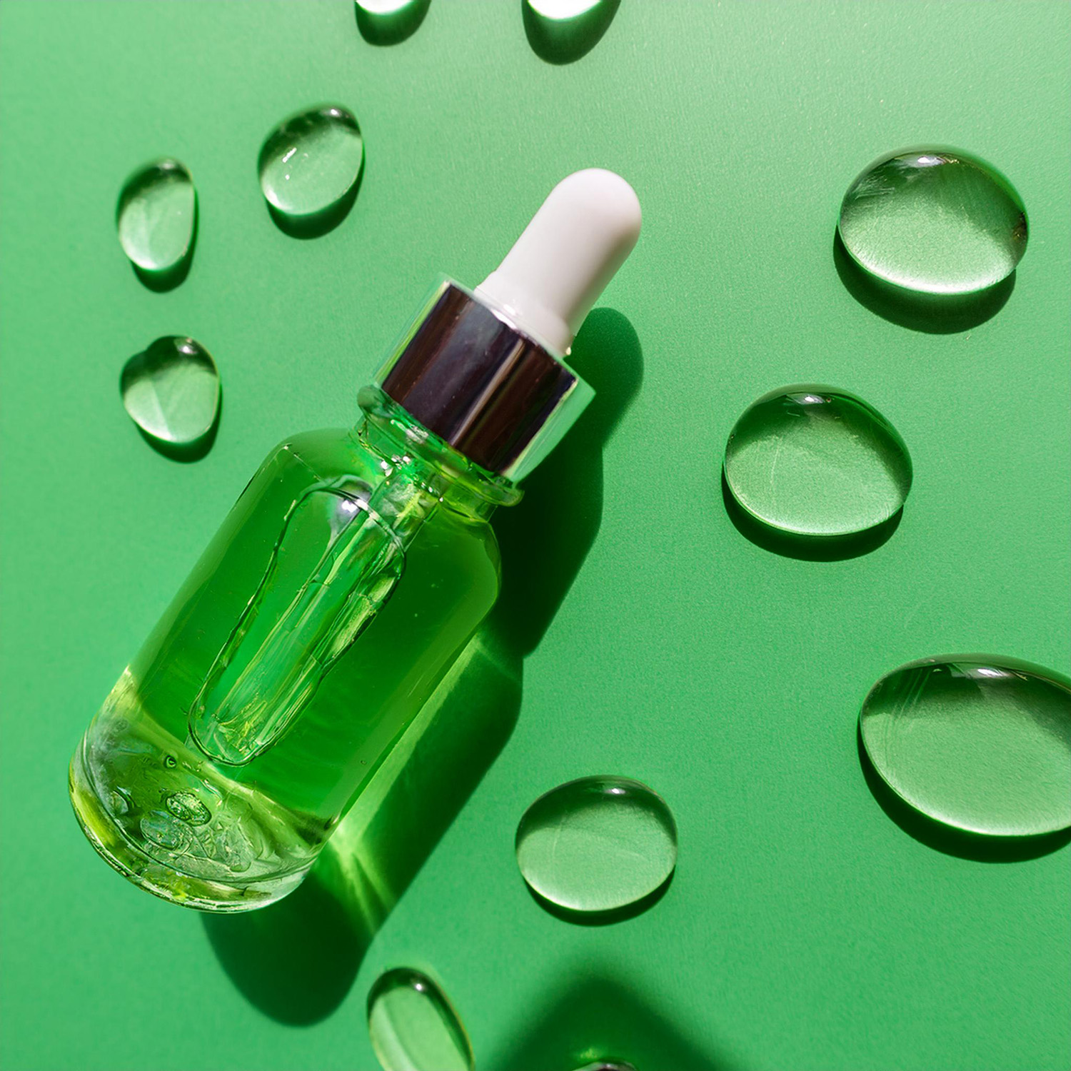 view of a glass bottle with pump closure full of transparent liquid – hyaluronic serum. Around the bottle with the serum there are several drops of spilled serum on the background. The background consists of a plain, light green background. The bottle with the serum is lying down. Top view and studio lighting